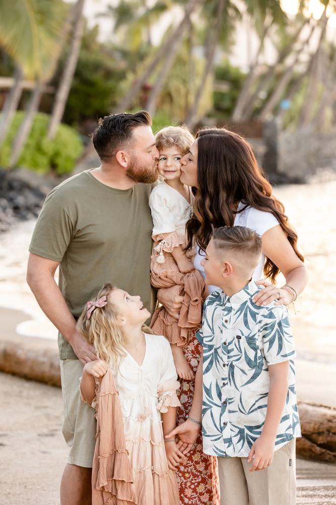 Family of five smiling on a beach, with parents kissing child's cheeks, children looking up happily.