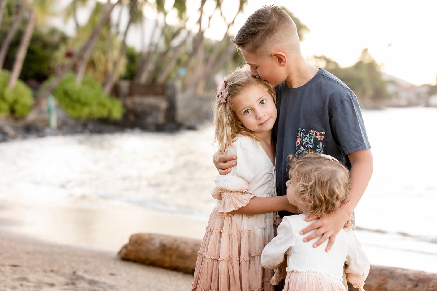 Three children hugging on a beach with palm trees in the background.
