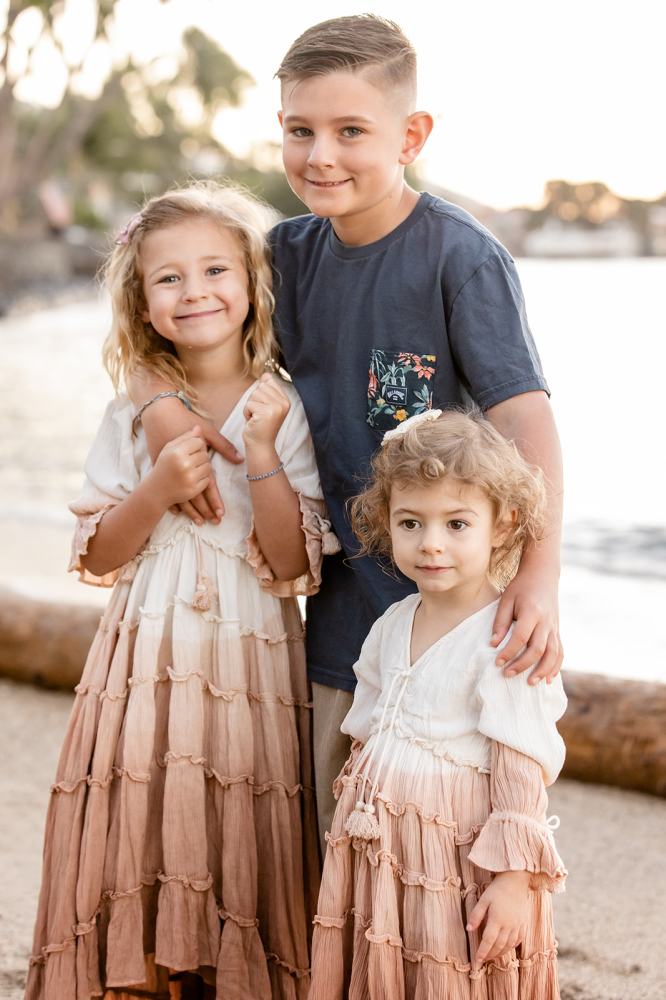 Three children smiling at the beach, with two girls in dresses and a boy in a blue shirt.