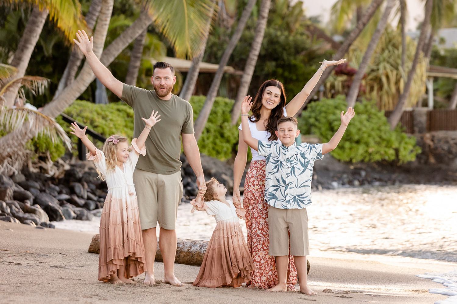 Smiling family of five posing with raised arms on a tropical beach with palm trees in the background.