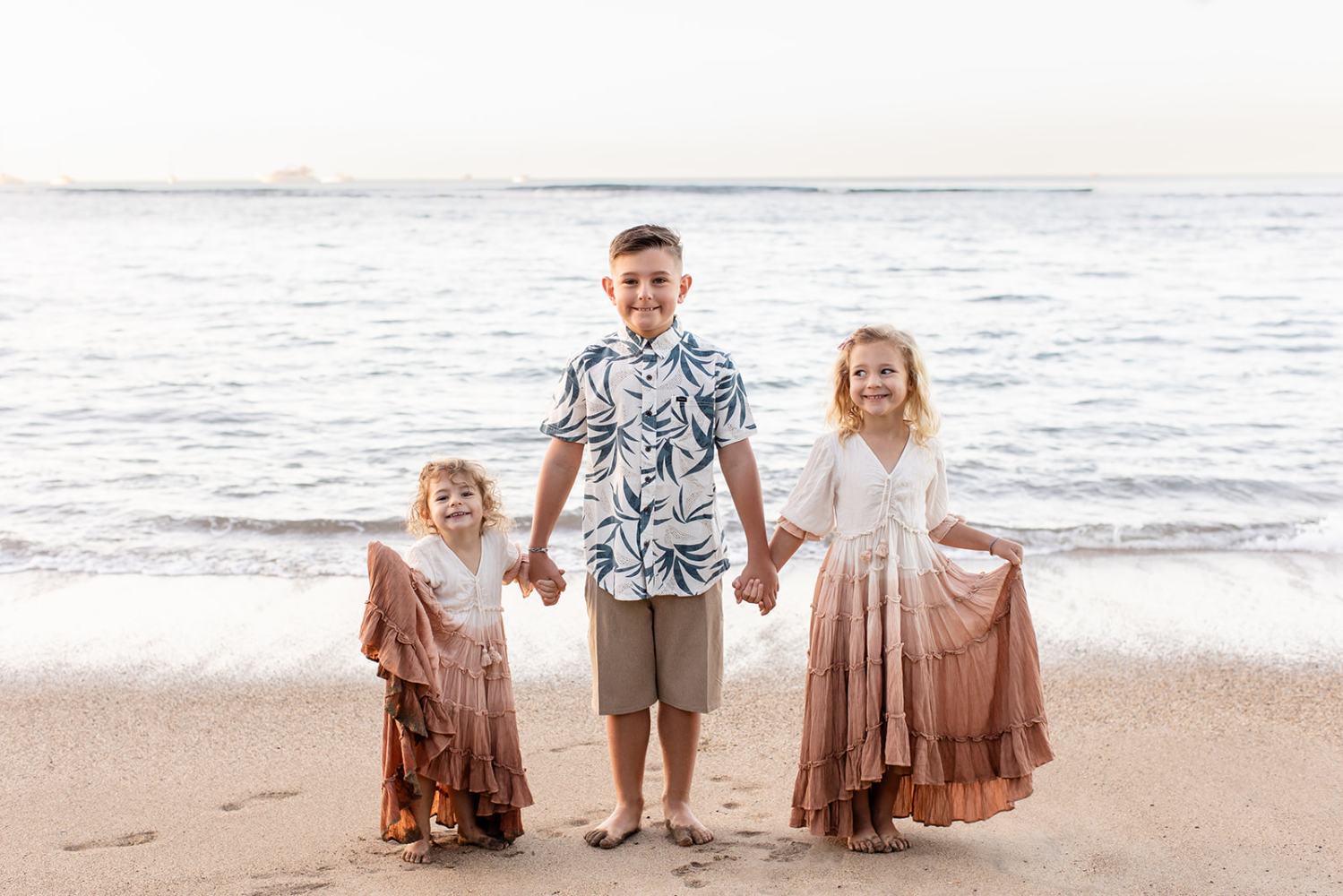 Three children holding hands, standing on a beach in front of the ocean.