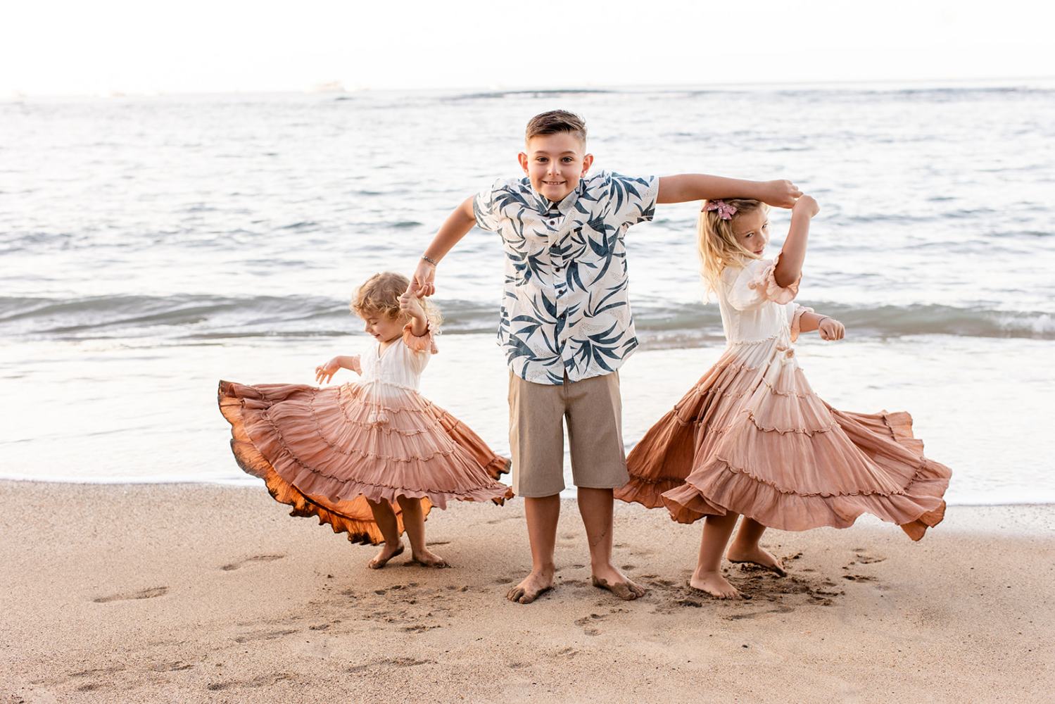Three children dancing on a beach; a boy in the center, two girls with dresses twirling beside him.
