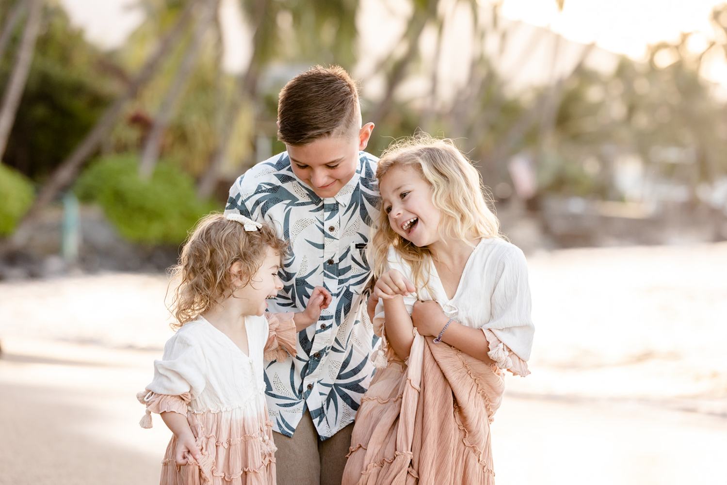 Three smiling children on a beach, surrounded by palm trees, wearing light-colored clothes.