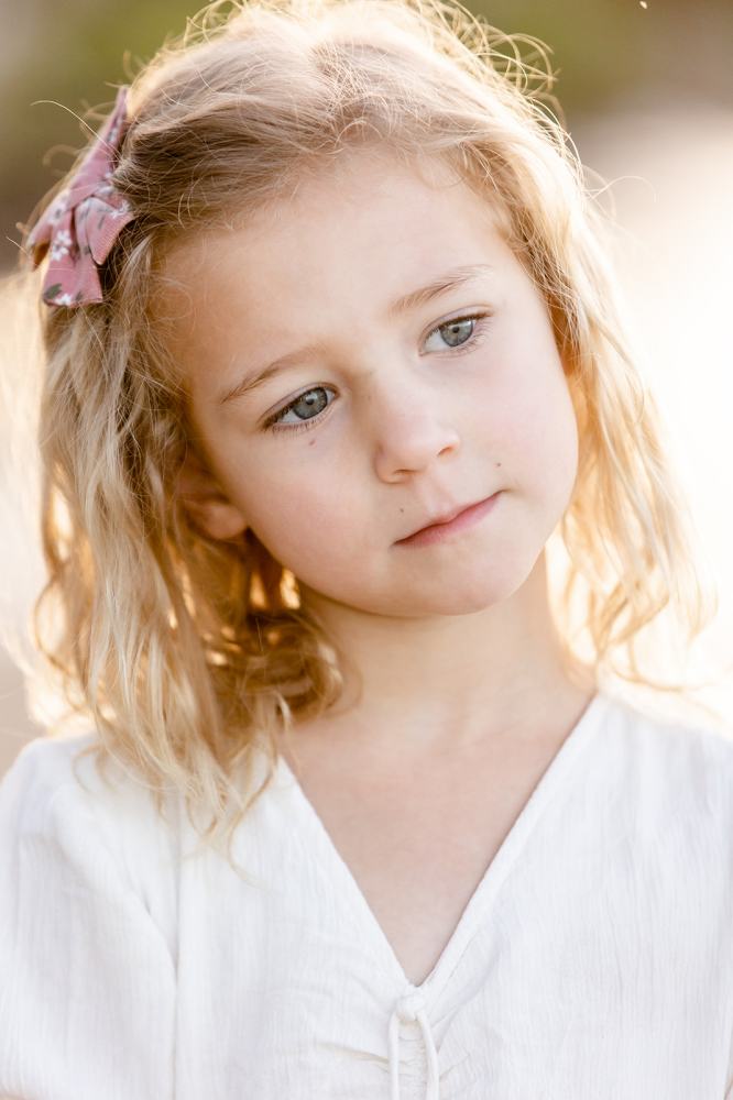 Young girl with blonde hair and pink bow looks thoughtful in soft lighting.