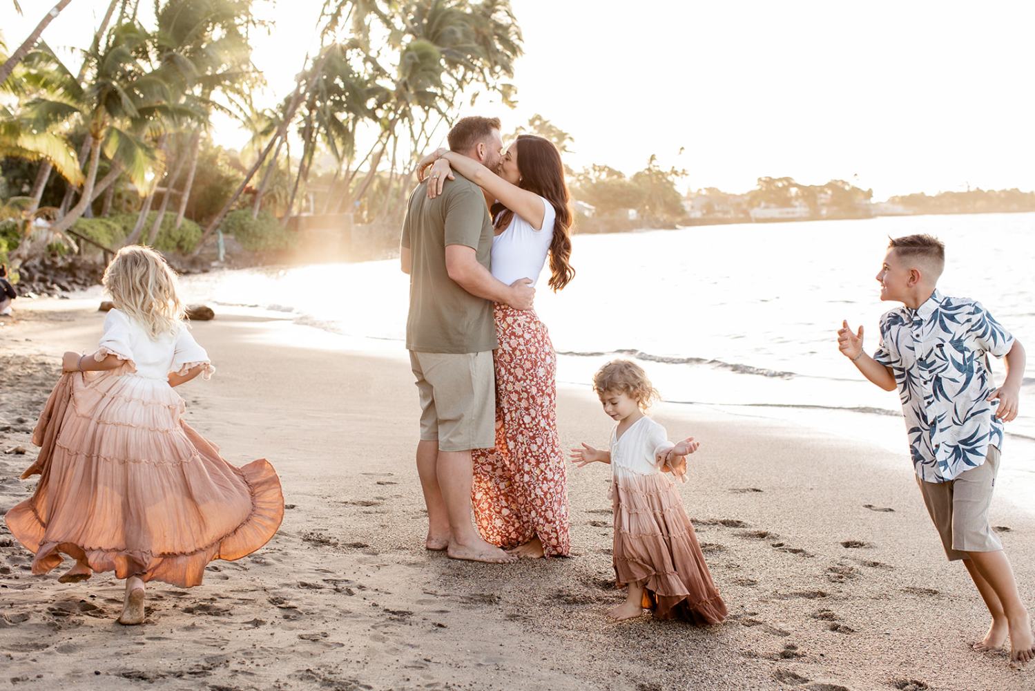 Family playing on a beach at sunset, palm trees in the background, adults hugging, children running.
