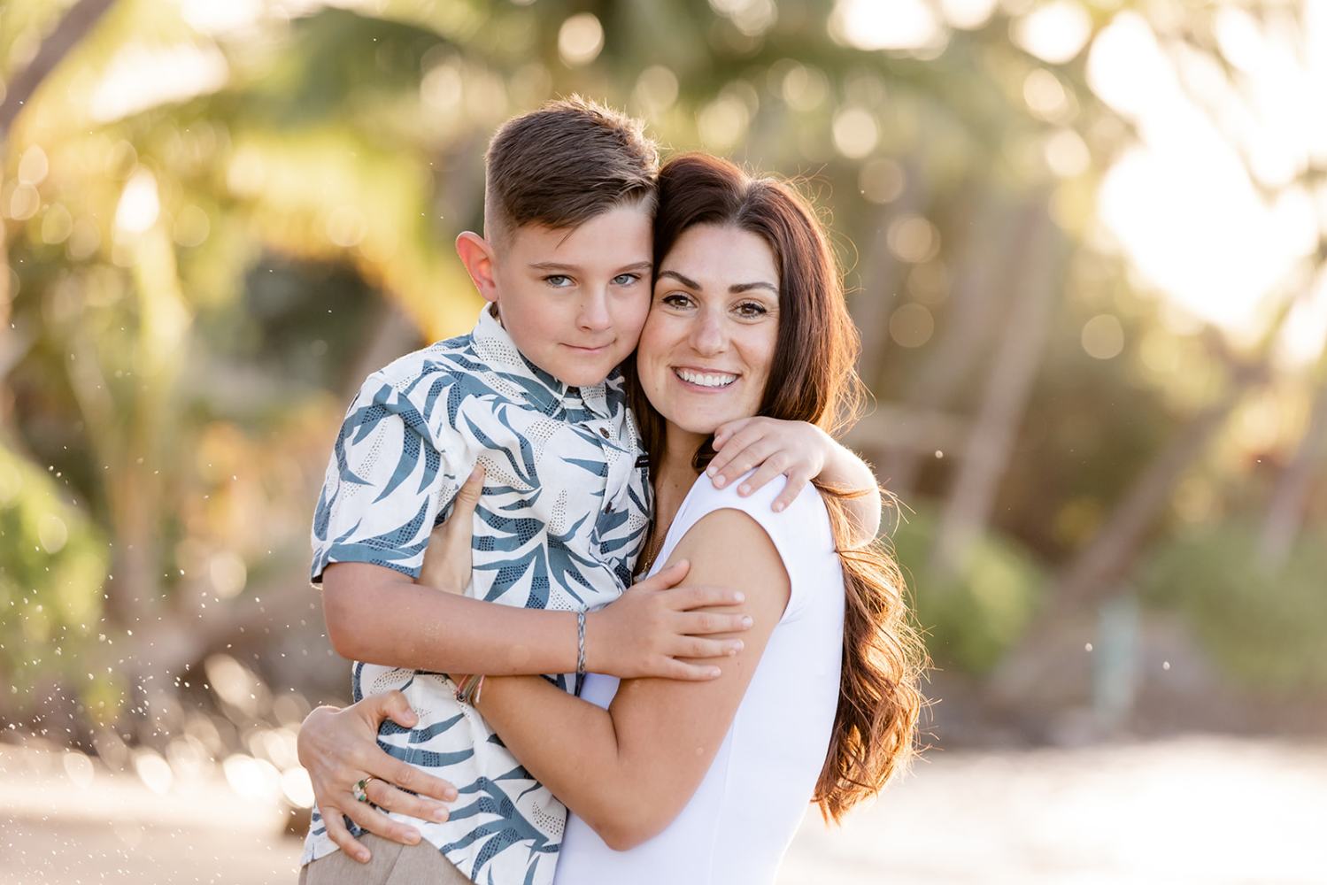Woman holding a boy on the beach with trees in the background.