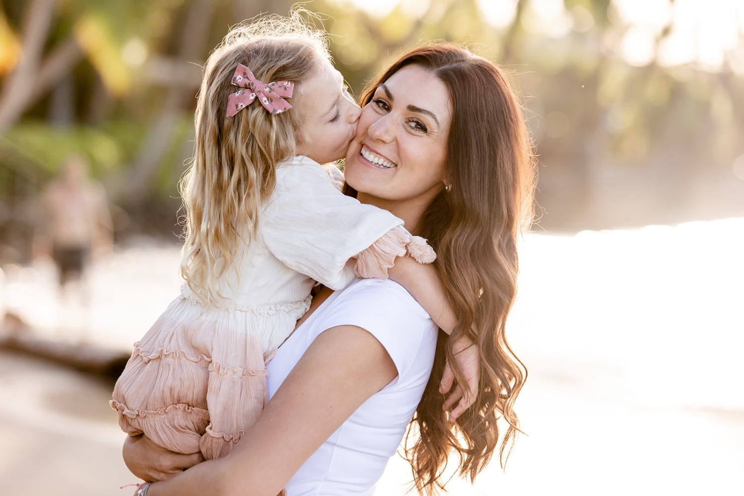 Woman holding a young girl kissing her cheek on a sunlit beach.