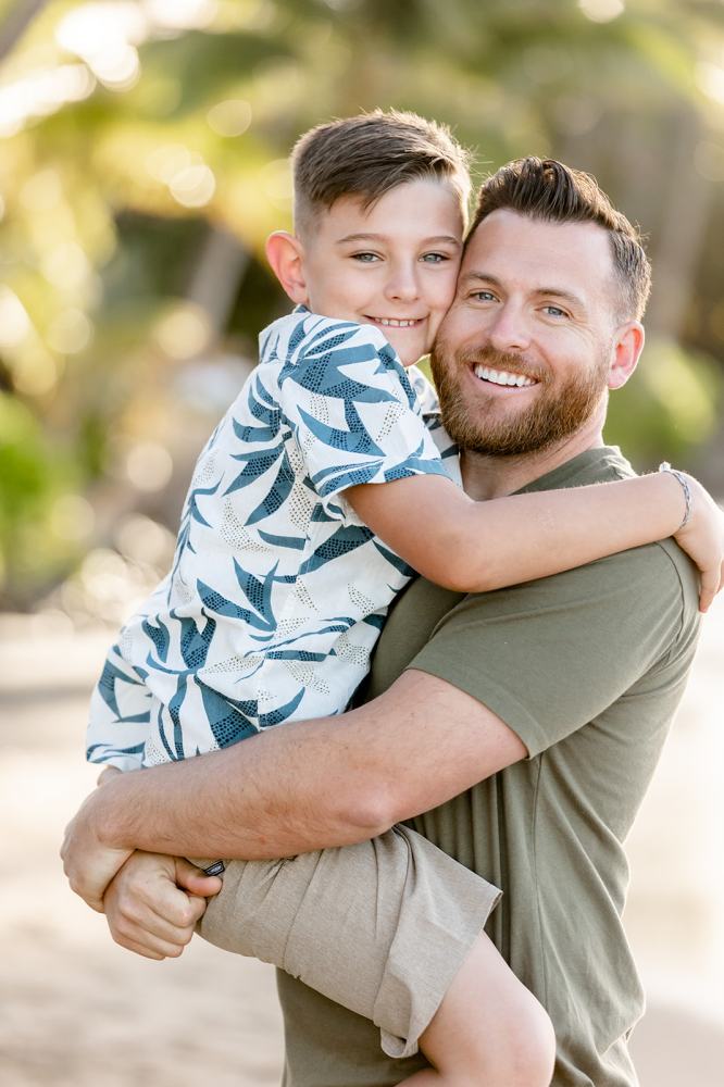 Man holding a smiling child in his arms at the beach with palm trees in the background.