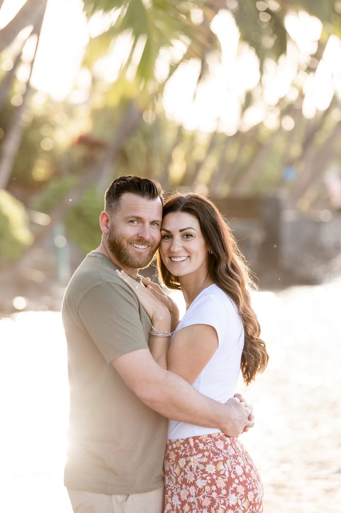 Couple embracing outdoors with palm trees and sunlight in the background.