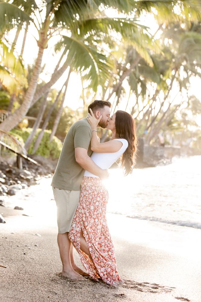 A couple kissing on a sunny beach with palm trees in the background.