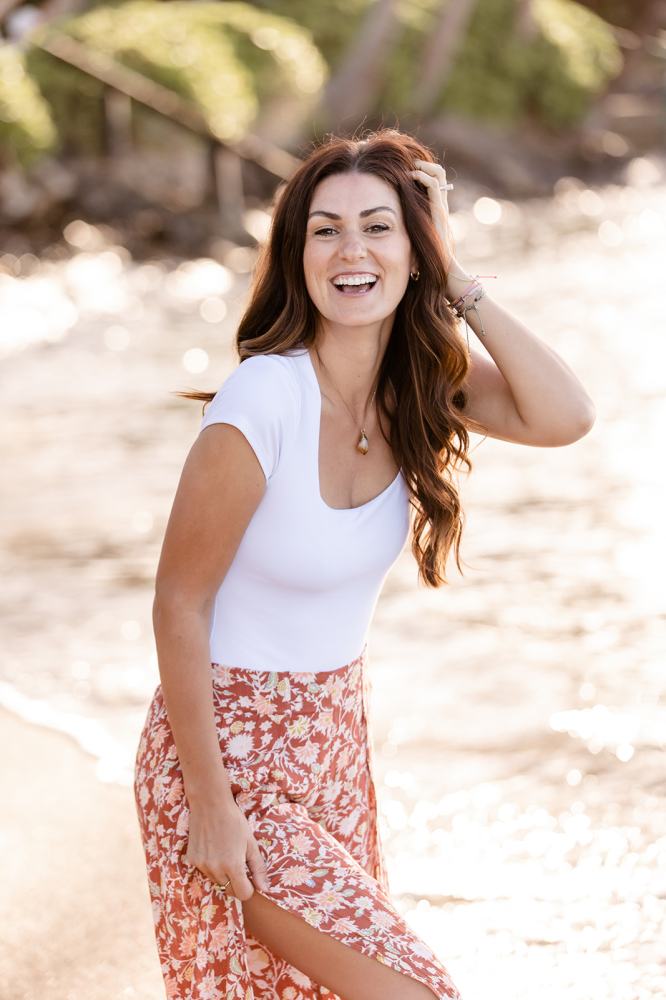 Woman smiling on beach, wearing white top and floral skirt, with hand in hair.