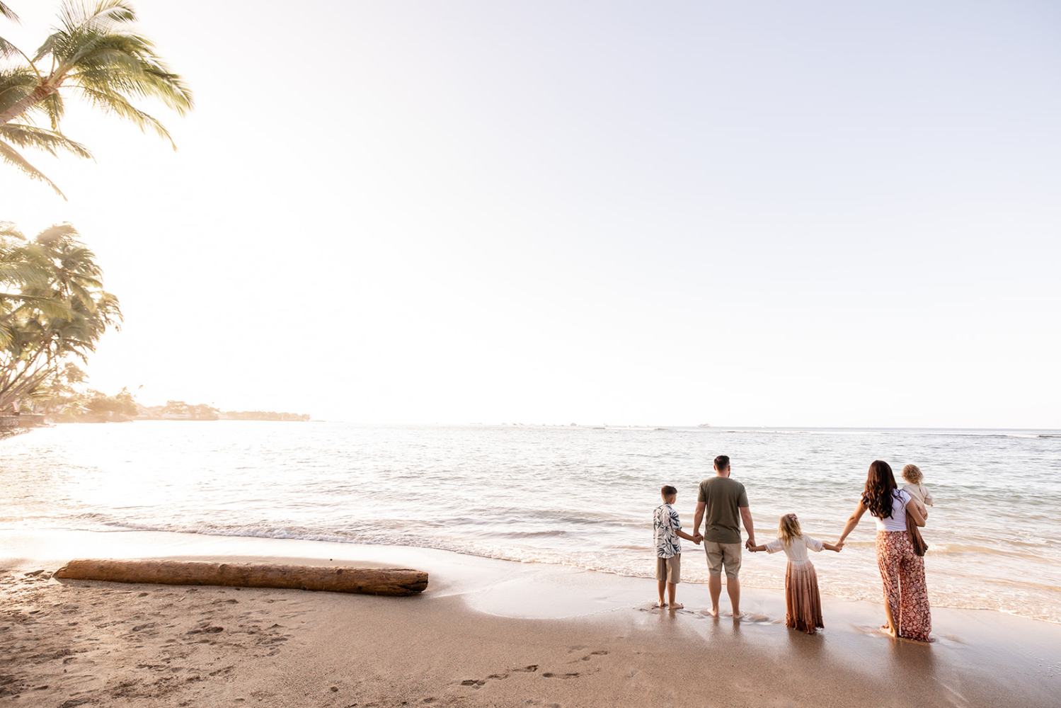 A family of five stands on a beach holding hands, facing the sea at sunset.