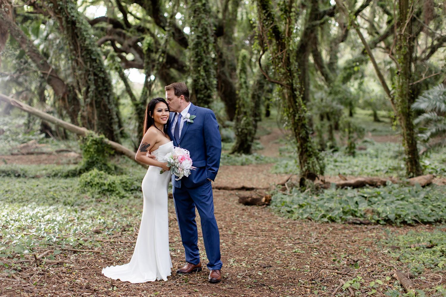 Couple in wedding attire standing in a lush, green forest path.
