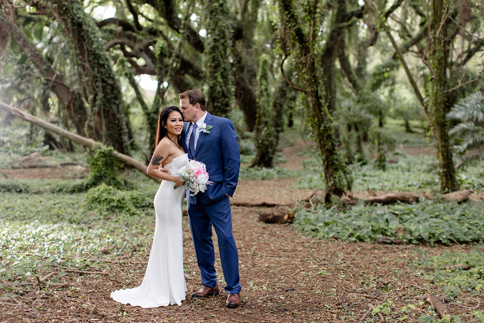 Couple in wedding attire standing in a lush, green forest path.