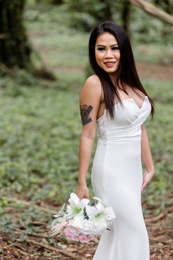 Woman in white dress holding flowers outdoors, showing tattoo on arm.