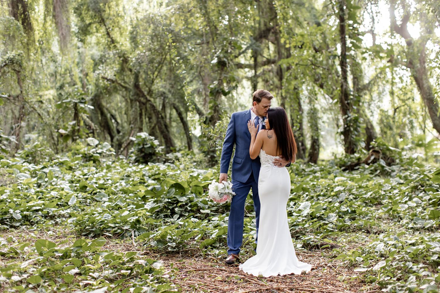 Couple in wedding attire embracing in lush green forest clearing.