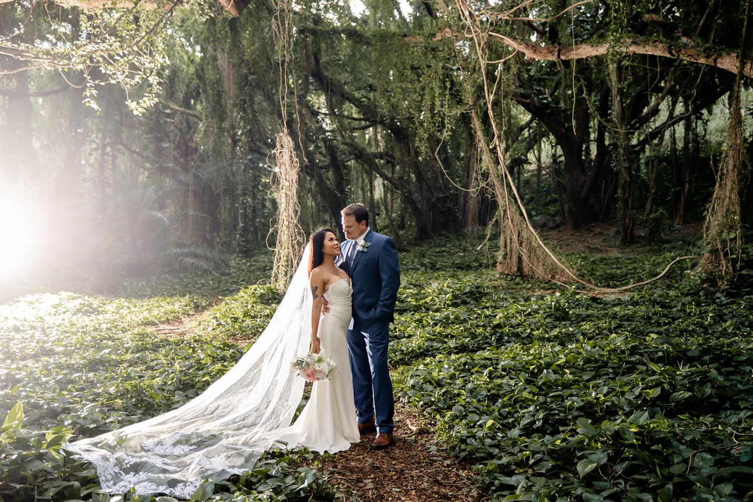 Bride and groom embracing in a lush forest with sunlight streaming through trees.