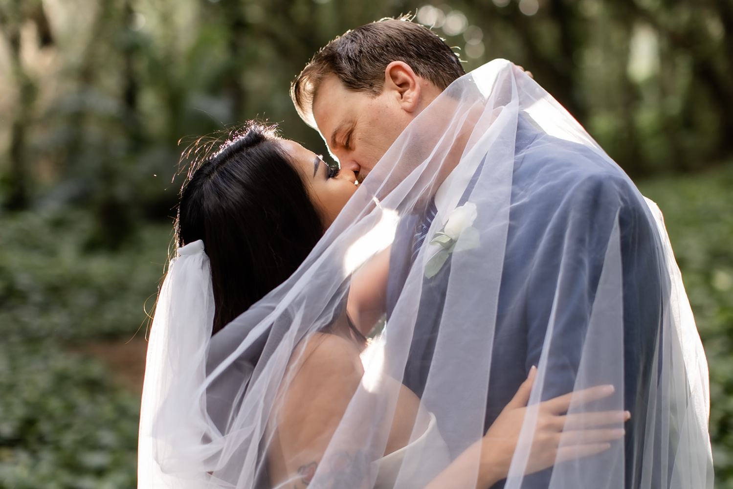 Bride and groom kissing under a veil in a forest setting.