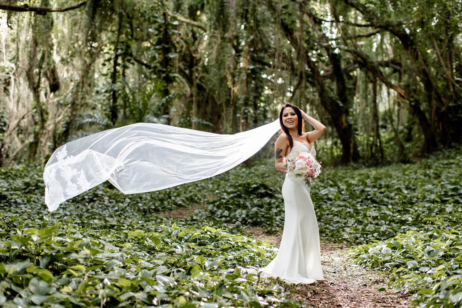 Bride in white dress with long veil stands in lush green forest, holding a bouquet, smiling.