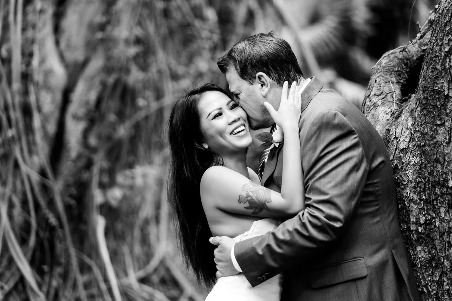 Smiling couple embracing and laughing in front of a forest backdrop in black and white.