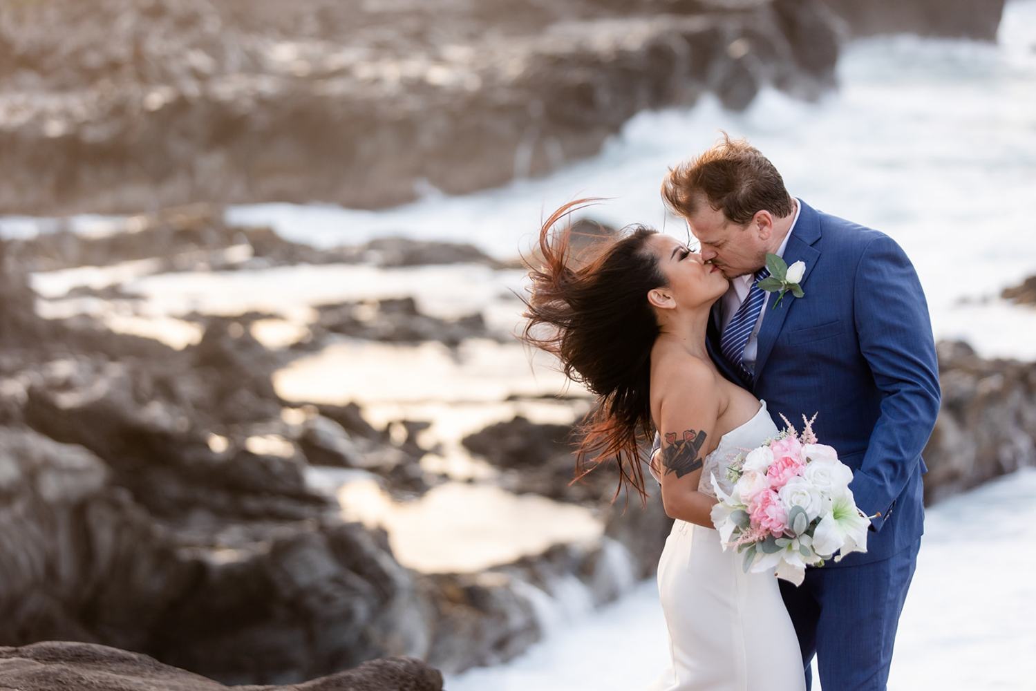 Couple in wedding attire kissing on rocky shoreline with ocean backdrop.
