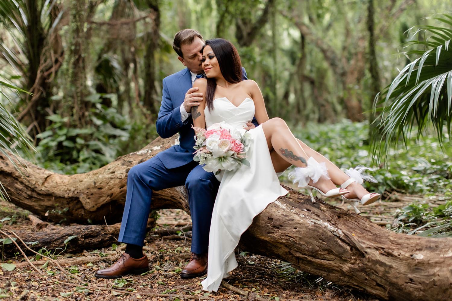 Couple in wedding attire sitting on a log in a forest, bride holding a bouquet.
