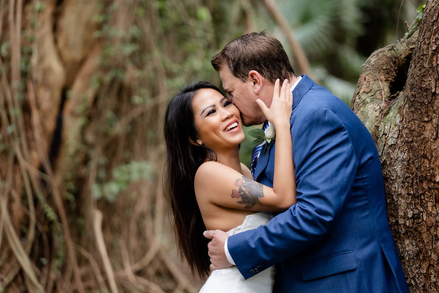 A couple embraces near a tree. The woman smiles while the man kisses her forehead.