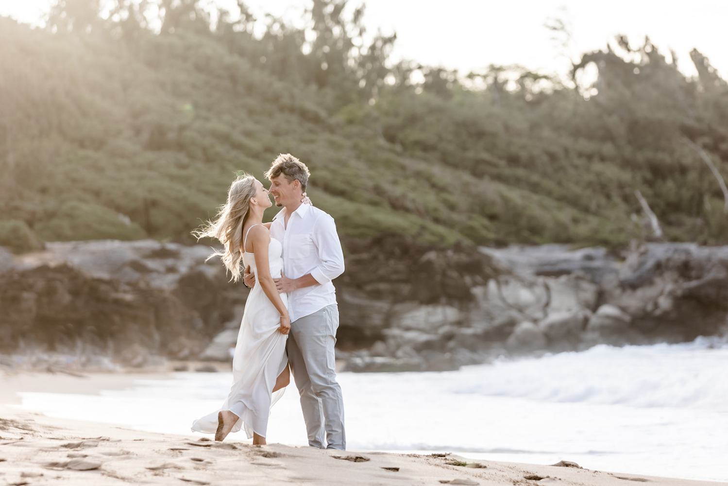 Couple embracing on a beach with waves and rocky cliffs in the background.