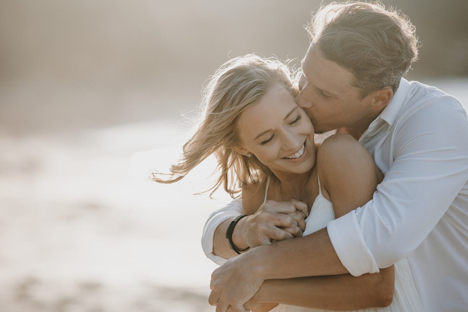 Couple embracing on a beach. Man kisses woman's cheek while she smiles.