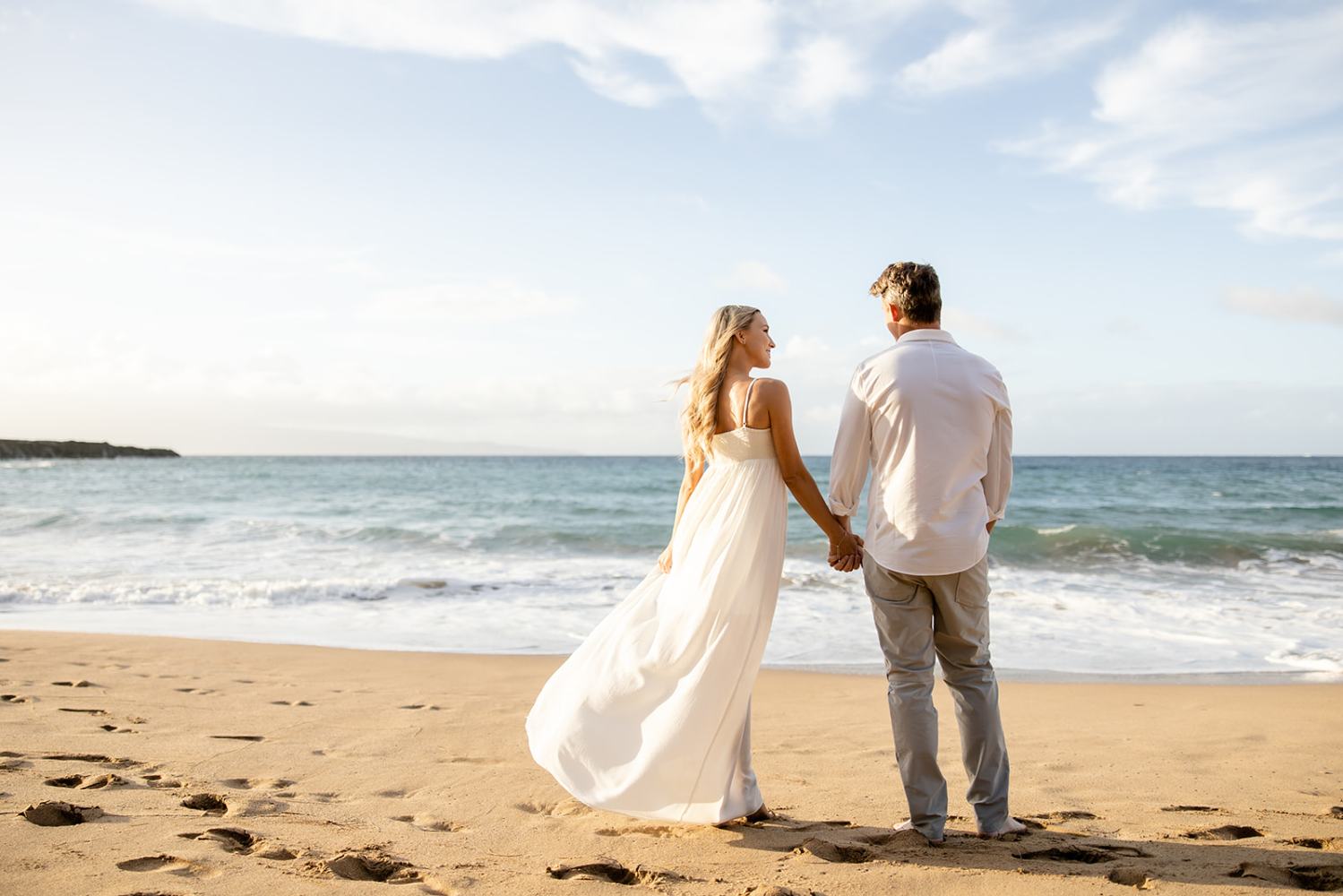 Couple holding hands on a beach, facing the ocean under a blue sky with scattered clouds.