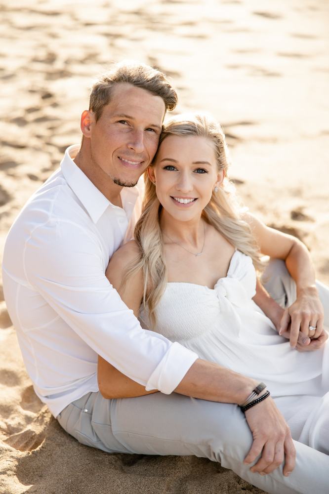 Couple in white attire sitting on beach sand, smiling at camera.