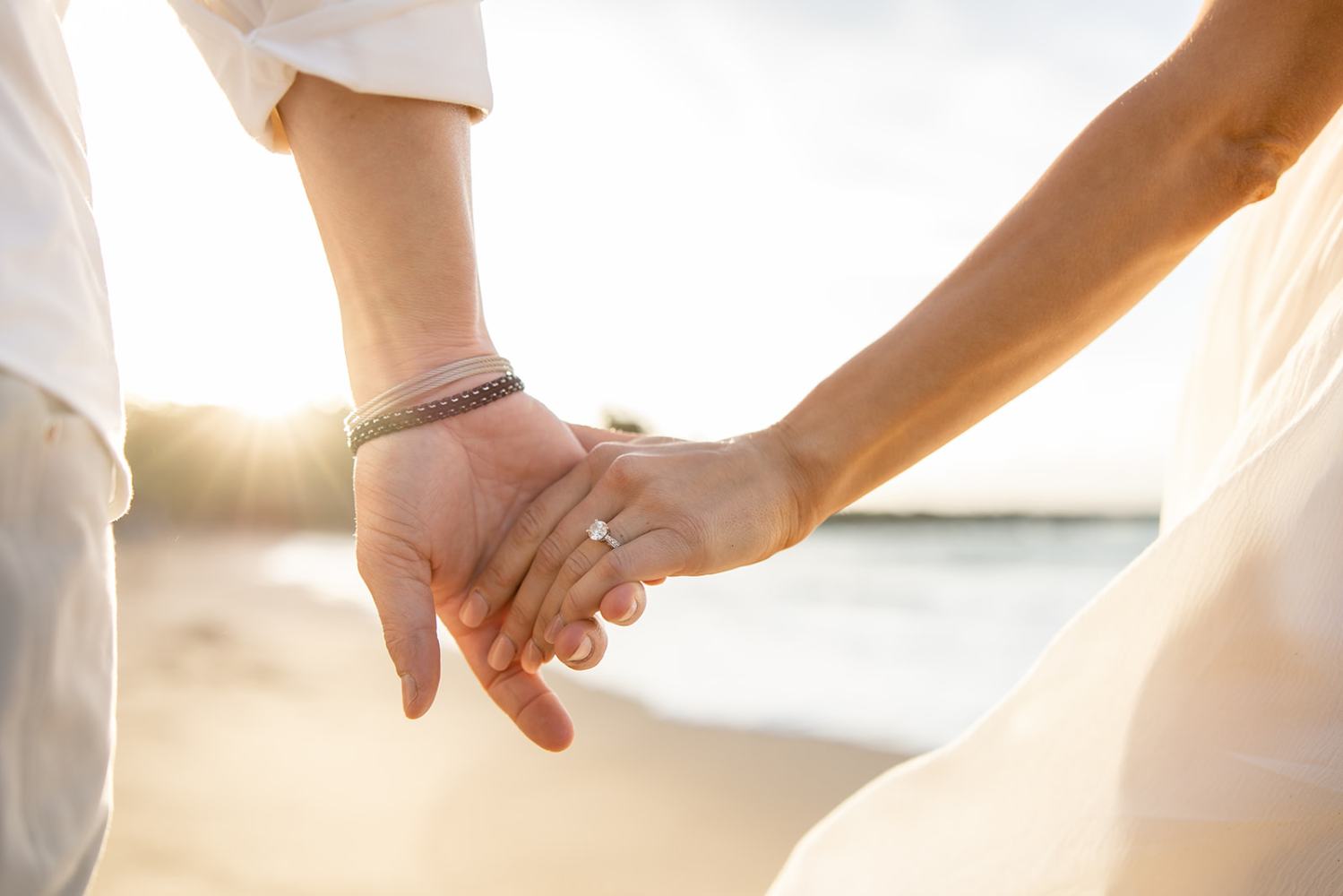 A couple holding hands on a beach, with a sunset in the background.