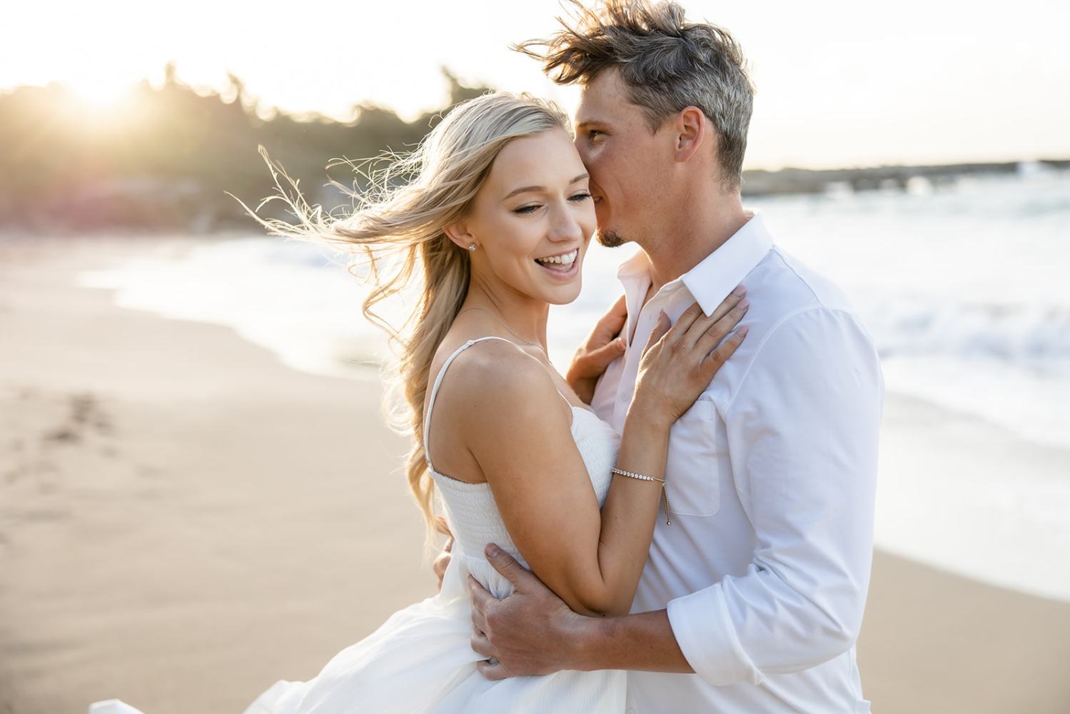 Man whispers to smiling woman on a beach at sunset, both in white attire.