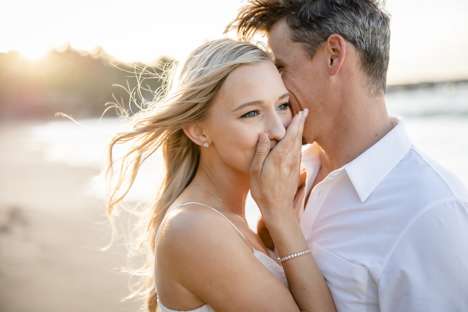 Smiling couple on a beach at sunset, woman covers mouth and looks surprised.