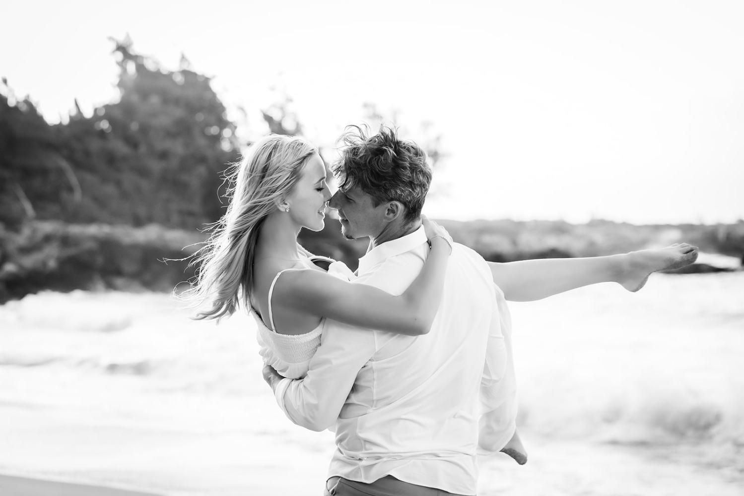 Man carrying woman on beach, both smiling, ocean waves in background.