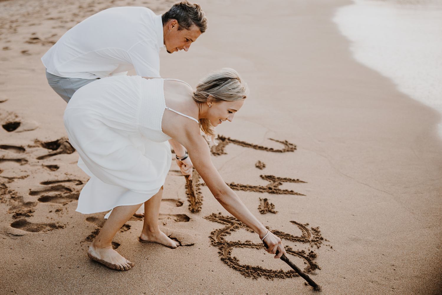 Couple writing in sand on beach, smiling, in casual white outfits.
