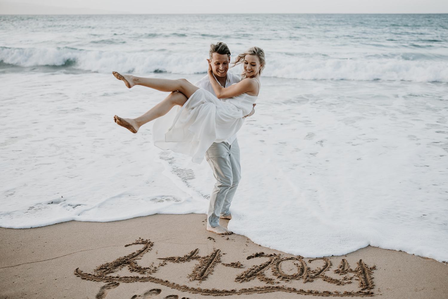 Couple on beach, person in white dress is carried, date '2-4-2024' written in sand.