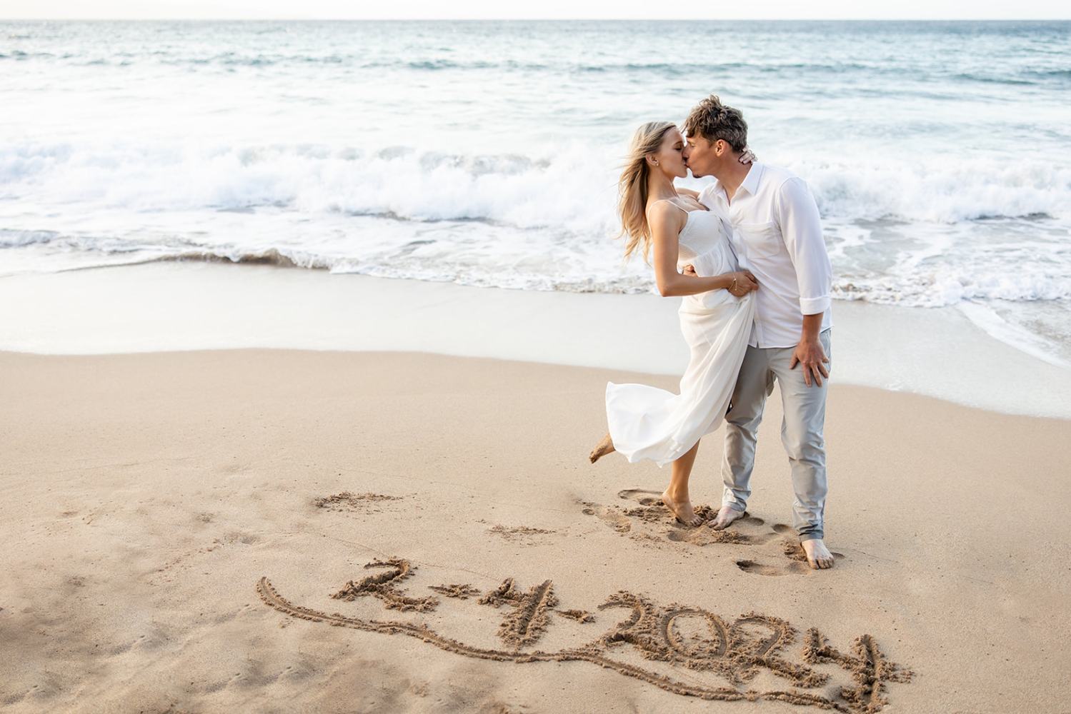 Couple kissing on beach with '2-4-2024' written in sand.