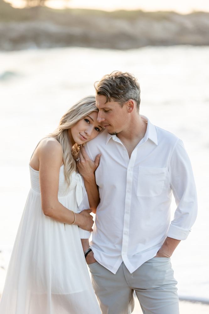 Couple in white clothing embracing by the beach, woman resting head on man's shoulder.