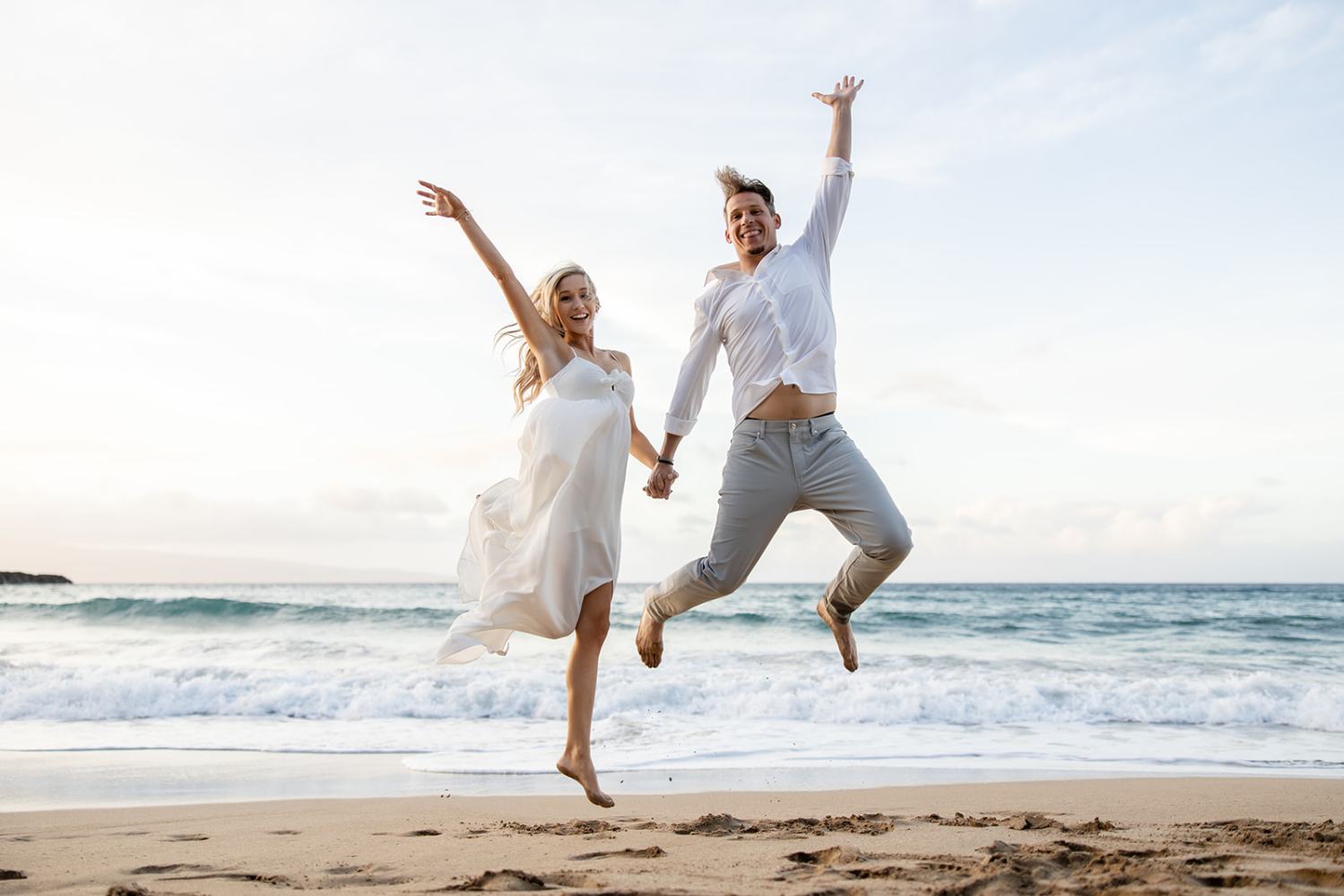 A couple in white jumping joyfully on a beach, holding hands near the ocean.