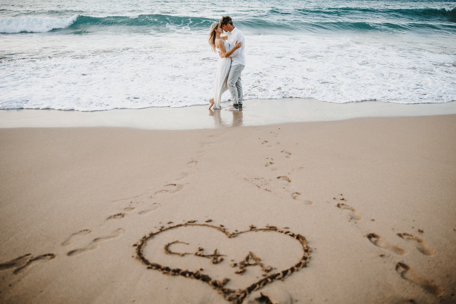Couple kissing on a beach near waves with initials in a heart drawn in the sand.