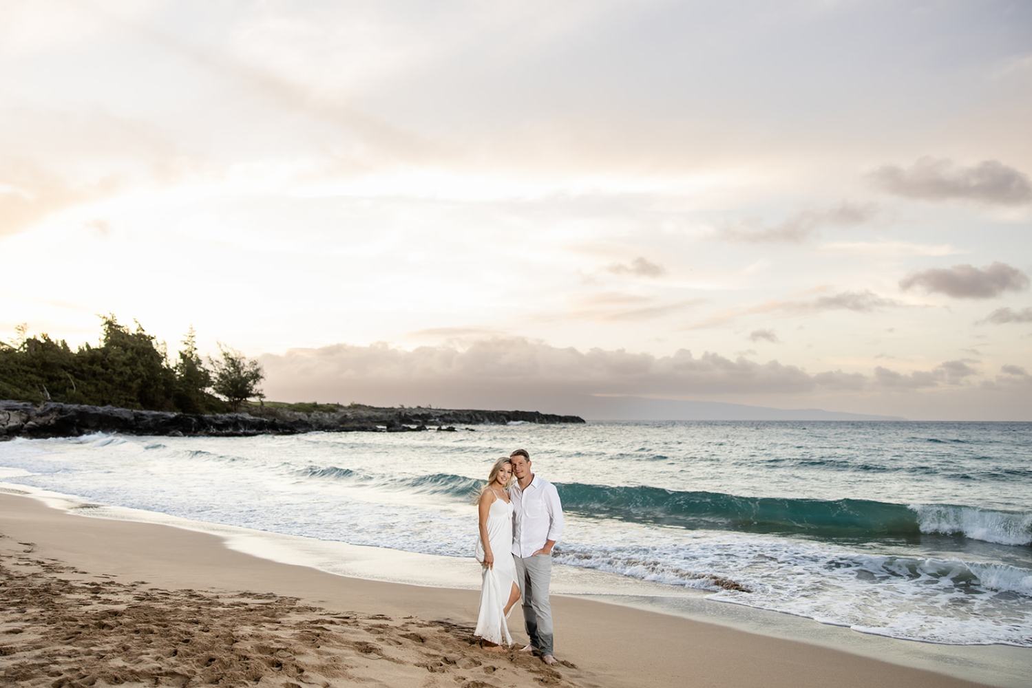 Couple standing on a sandy beach by the ocean during sunset.
