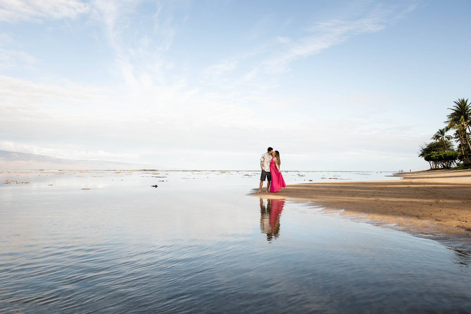 Couple stands on a beach, with reflections in the shallow water, under a blue sky with clouds.