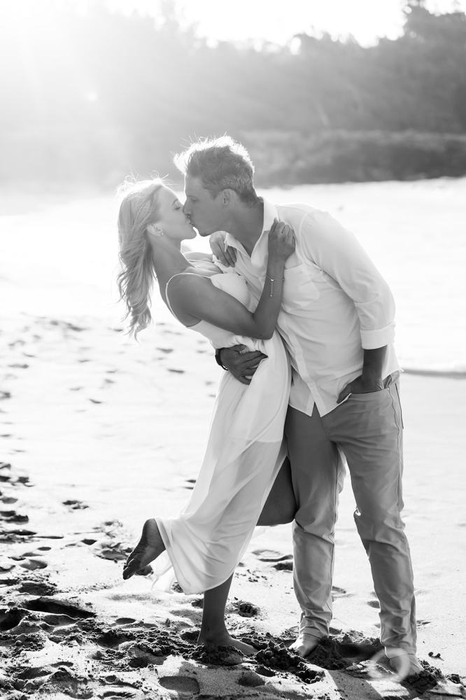 Black and white photo of a couple kissing on a sandy beach with sunlight in the background.