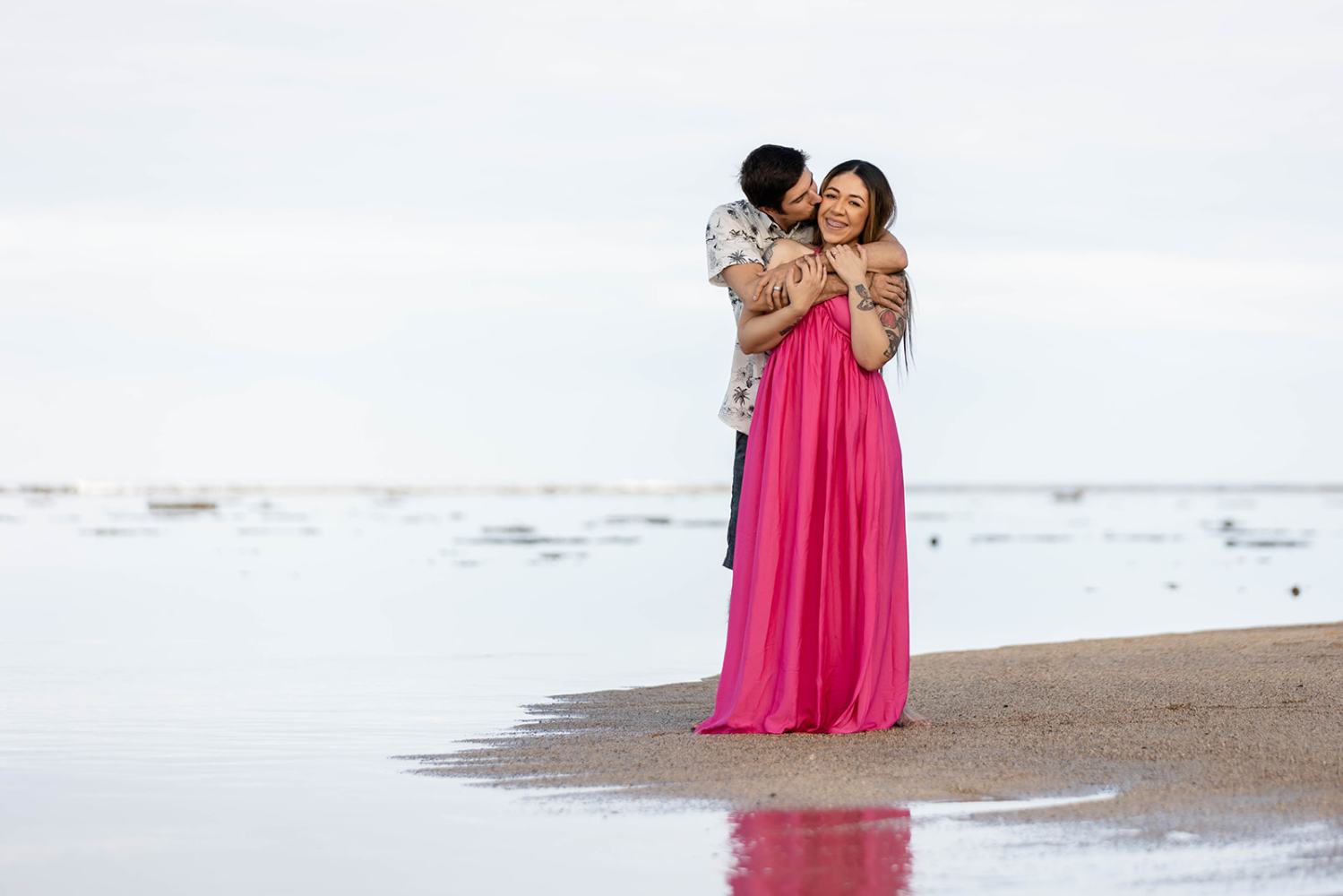 Couple embraces on a sandy beach, woman in a pink dress, man in a patterned shirt.