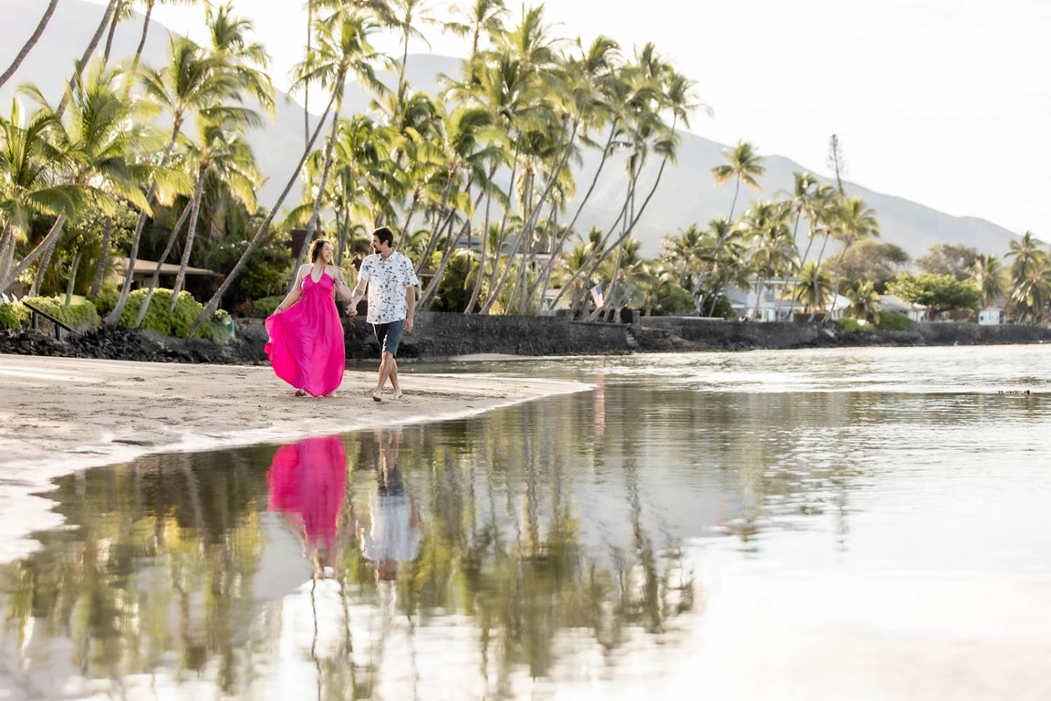 Couple walking on a tropical beach with palm trees, woman in pink dress.