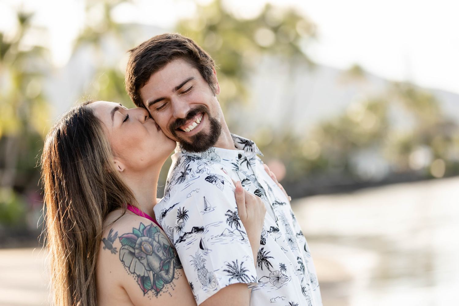 Woman kissing man on cheek by the beach, both smiling.