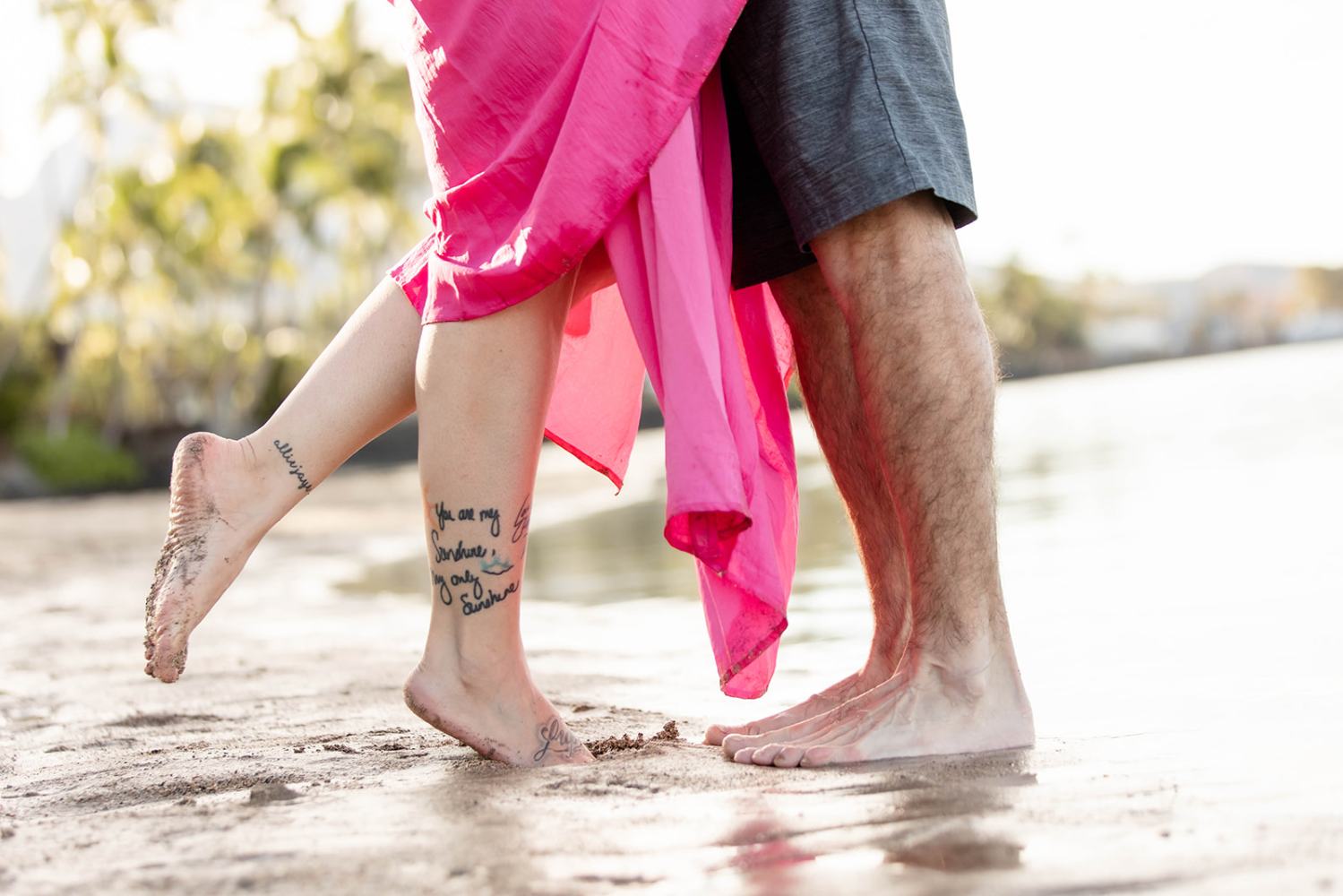 Couple standing barefoot on a beach, woman's pink dress and tattoos visible.