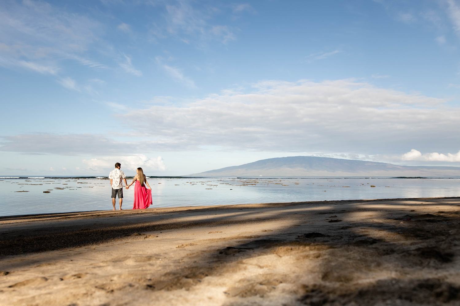 Couple holding hands on a sandy beach with mountains in the background under a blue sky.
