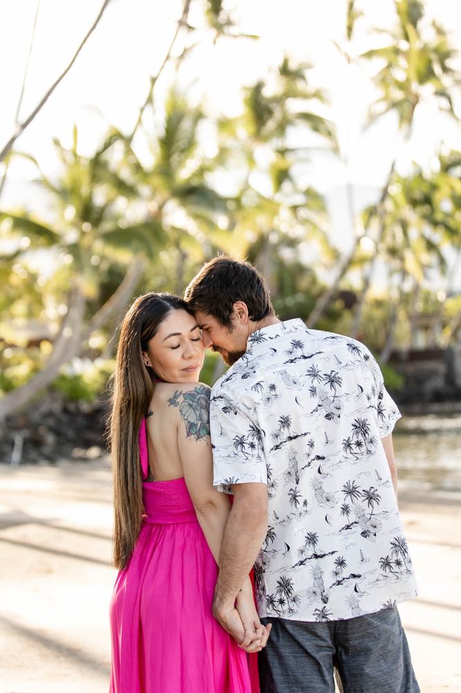 Couple holding hands, facing each other on a sunny beach with palm trees.
